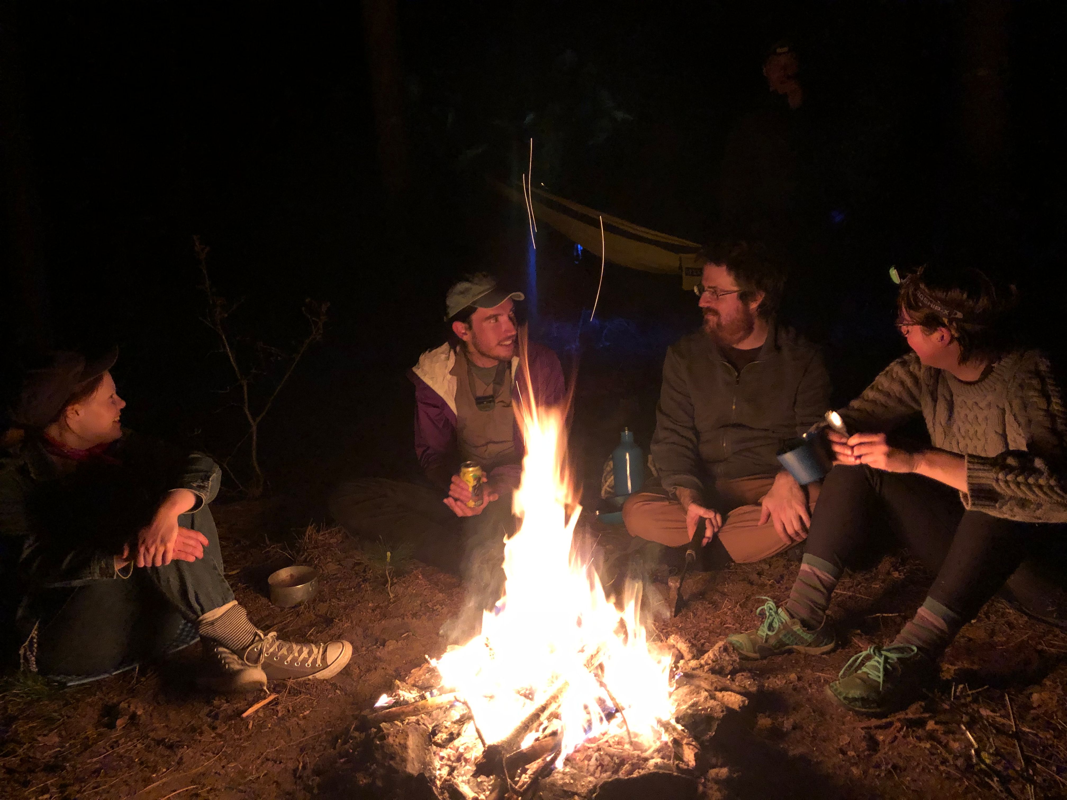 Summiteers sit around a camp bonfire in the woods. (Circa spring 2020 beginning of Covid)